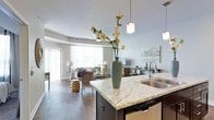 This single-floor residence is designed with an open-concept layout that seamlessly connects the kitchen, dining, and living areas into one cohesive, flowing space. The kitchen serves as a central hub, anchored by a substantial island topped with striking granite countertops featuring grey and white veining. A stainless steel sink is integrated into the island, which is finished with a dark base, creating a bold contrast against the light stone surface. Pendant lighting hangs above, providing focused illumination and adding a contemporary design element.

Dark hardwood flooring extends throughout the space, grounding the interior with warmth and visual continuity. The living area benefits from generous natural light streaming through large windows dressed with sheer curtains and blinds, creating a bright and welcoming atmosphere. Neutral grey walls provide a versatile backdrop throughout. A ceiling fan is installed in the living area, contributing to both comfort and function. The dining area is positioned between the kitchen and living room, reinforcing the open, connected feel of the overall layout.