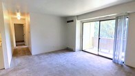 This single-floor space features a clean, minimalist design with a focus on functionality and natural light. The main living area is finished with wall-to-wall carpeting in a neutral tone, providing a soft and comfortable foundation throughout the room. Smooth white walls and a flat white ceiling create a bright, airy atmosphere that feels open and easy to personalize.

A standout feature of this space is the full-width sliding glass door, which floods the interior with abundant natural light and offers direct access to a private outdoor balcony. Through the glass, views of mature trees and greenery add a natural, calming element to the interior ambiance. The balcony itself features wooden railing detailing, adding a warm contrast to the otherwise neutral interior palette.

Practical infrastructure is well integrated, with a ceiling-mounted HVAC vent and baseboard heating ensuring year-round climate comfort. Electrical outlets are conveniently positioned along the walls. The overall layout is simple and versatile, offering a blank canvas well-suited for a variety of furniture arrangements and interior styles.