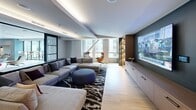 This stylish, contemporary living room offers a well-appointed and thoughtfully designed lounge space. The room is anchored by a generous L-shaped sectional sofa in light gray, positioned atop a bold black-and-white abstract area rug, and complemented by a round blue ottoman. A pair of designer accent chairs — one orange, one gray — are positioned near the large windows, creating an inviting reading nook bathed in natural light with views of the surrounding cityscape.

The entertainment wall features a large wall-mounted flat-screen TV set against a dark accent wall, flanked by warm wood built-in shelving with integrated lighting, offering ample display and storage space. On the opposite side of the room, a built-in wet bar is showcased with a striking blue mosaic tile backsplash and warm wood cabinetry, adding both function and visual interest.

Light wood-look flooring runs throughout, contributing to a clean and modern aesthetic. The ceiling incorporates cove lighting that casts a warm ambient glow, adding to the room's polished atmosphere. Large black-framed windows allow natural light to flood the space. Sliding glass-panel doors with frosted detailing provide separation from adjacent areas while maintaining an open, connected feel.