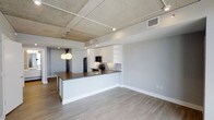 This 2-bedroom, 2-bathroom unit offers a well-designed open-concept layout that seamlessly connects the living and kitchen areas. The kitchen is a standout feature, showcasing white cabinetry with open shelving, dark countertops, a subway tile backsplash, and black stainless steel appliances including a refrigerator and range. A peninsula counter provides additional prep space and doubles as a casual dining or entertaining area, complemented by modern globe pendant lighting overhead. The ceiling features an exposed concrete finish with visible track lighting and HVAC infrastructure, lending the space an industrial-chic character that contrasts attractively with the clean, light gray walls. Light-toned hardwood-style flooring runs throughout, adding warmth and visual continuity across the open floor plan. Natural light filters through the space, enhancing its airy and modern ambiance. The overall design balances contemporary aesthetics with functional living, making it a practical and visually appealing home. With two bedrooms and two full bathrooms, the layout offers comfortable accommodations suited for a variety of living arrangements.