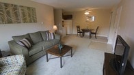 This single-floor residential space offers a practical and cohesive open-concept layout that seamlessly connects the living, dining, and kitchen areas into one unified living environment. The living area is anchored by plush wall-to-wall carpeting in a neutral tone, creating a warm and inviting atmosphere. Beige walls provide a clean, understated backdrop that enhances the sense of openness throughout the space. The dining area is positioned centrally within the open floor plan, featuring a circular dining table with cushioned chairs and a ceiling fan with a light fixture overhead, offering both comfort and functionality. Adjacent to the dining space, the kitchen is efficiently laid out with dark cabinetry, a white refrigerator, and tile flooring that transitions naturally from the carpeted living areas. A framed mirror on the dining room wall adds depth and reflects natural light into the space. Decorative wall art in the living area adds a subtle natural touch, complementing the overall earthy color palette. The single-floor layout ensures easy accessibility and a smooth flow between all areas, making this a well-designed, functional living space suited for everyday comfort.