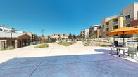 This expansive outdoor amenity area serves as the central hub of a modern residential community, thoughtfully designed to encourage recreation and relaxation. The grounds are framed by multi-story residential buildings featuring earthy stucco exteriors in warm terracotta and tan tones, with private balconies overlooking the shared spaces below.

Meandering concrete walkways connect the various amenity zones, bordered by a carefully curated xeriscape landscape that blends ornamental grasses, native shrubs, decorative boulders, and gravel beds with patches of lush green lawn. This water-conscious design reflects a Southwest-inspired aesthetic well-suited to the arid climate.

A standout feature is the dedicated putting green, nestled alongside a dramatic boulder retaining wall that adds natural texture and visual interest to the grounds. Adjacent to the green, a covered pergola area provides shaded outdoor seating, ideal for gathering and leisure. The community clubhouse anchors one end of the property, presenting a welcoming stucco façade with large windows, an inviting entrance walkway, and an adjacent patio furnished with tables and colorful umbrellas. A gated pool area rounds out the recreational offerings, completing this resort-style outdoor environment. Tall lamp posts line the pathways, ensuring the space remains functional and inviting throughout the day.