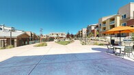 This expansive outdoor amenity area serves as the central hub of a modern residential community, thoughtfully designed to encourage recreation and relaxation. The grounds are framed by multi-story residential buildings featuring earthy stucco exteriors in warm terracotta and tan tones, with private balconies overlooking the shared spaces below.

Meandering concrete walkways connect the various amenity zones, bordered by a carefully curated xeriscape landscape that blends ornamental grasses, native shrubs, decorative boulders, and gravel beds with patches of lush green lawn. This water-conscious design reflects a Southwest-inspired aesthetic well-suited to the arid climate.

A standout feature is the dedicated putting green, nestled alongside a dramatic boulder retaining wall that adds natural texture and visual interest to the grounds. Adjacent to the green, a covered pergola area provides shaded outdoor seating, ideal for gathering and leisure. The community clubhouse anchors one end of the property, presenting a welcoming stucco façade with large windows, an inviting entrance walkway, and an adjacent patio furnished with tables and colorful umbrellas. A gated pool area rounds out the recreational offerings, completing this resort-style outdoor environment. Tall lamp posts line the pathways, ensuring the space remains functional and inviting throughout the day.
