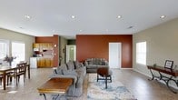 This single-floor space is designed with an open-concept layout that seamlessly connects the living area to the kitchen, creating a sense of spaciousness and ease of movement. The living room is a focal point of the home, anchored by a large sectional sofa arranged around a patterned area rug and complemented by a round wooden coffee table and a rectangular wooden side table. Bold terracotta-toned accent walls add warmth and visual depth to the space, contrasting with the neutral beige tones of the remaining walls and the light-toned tile flooring that runs throughout. Recessed ceiling lights provide consistent overhead illumination across the open space. A white paneled door is visible along the accent wall, offering access to an adjoining area. Natural light filters in through a window positioned on the side wall. The kitchen, visible in the background, features warm wood-toned cabinetry and continues the cohesive color palette established in the living area. The overall ambiance of the space is casual and inviting, with a practical layout suited for everyday living.