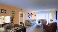 This single-floor residence offers a welcoming and functional layout designed for comfortable everyday living. The living room serves as the heart of the space, featuring warm peach-toned accent walls that create an inviting atmosphere. Plush wall-to-wall carpeting adds a sense of coziness underfoot, while a large window dressed with vertical blinds allows natural light to fill the room. The living area is spacious enough to accommodate a full sofa and loveseat arrangement centered around a glass-top coffee table, making it ideal for both relaxing and entertaining. Decorative wall-mounted shelving adds a stylish, modern touch to the living space. A dining area sits adjacent to the living room, separated subtly by the room's natural flow rather than any hard partition, creating an open and connected feel. The dining space features a dark wood table with matching chairs, complemented by a decorative vase with tall dried botanicals. A hallway visible from the living area leads to additional rooms, suggesting a thoughtful and accessible floor plan. The textured ceiling and neutral trim throughout provide a clean, cohesive backdrop that ties all the elements of this single-level home together.