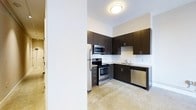 This single-floor space showcases a thoughtfully designed interior with a strong emphasis on modern aesthetics. The kitchen is a standout feature, outfitted with rich dark-espresso cabinetry that extends to the ceiling, providing generous storage space. A full suite of stainless steel appliances — including a refrigerator, electric range, built-in microwave, and dishwasher — adds a polished, professional feel to the space. Stone-look countertops complement the cabinetry, while a classic white subway tile backsplash introduces a clean, timeless contrast. The flooring throughout features a light-toned, speckled finish that enhances the sense of openness and reflects the ambient light well. Flush-mount ceiling lighting provides consistent illumination across the kitchen area. The layout is efficient and functional, with an L-shaped kitchen configuration that maximizes workspace. A structural column near the kitchen entry adds an architectural element to the design, subtly defining the transition between spaces. The overall ambiance is contemporary and clean, offering a well-appointed environment suited for modern living.