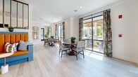 Located at 4029 Flat Shoals Pkwy in Decatur, Georgia, this single-floor space offers a thoughtfully designed dining and kitchen area that blends functionality with contemporary style. The open-concept layout is anchored by light wood-tone flooring that runs throughout, contributing to a clean, airy aesthetic.

The dining area accommodates multiple round tables surrounded by upholstered chairs, creating an inviting setting for shared meals or social gatherings. Oversized black-framed windows line the walls, flooding the space with abundant natural light and offering pleasant views of the surrounding greenery and outdoor covered areas. Bold floral drapery panels add a vibrant, decorative contrast to the neutral white walls.

The kitchen features a striking blue island with distinctive X-panel detailing and a crisp white countertop, complemented by a row of quilted blue barstools with brass-toned legs. Three large copper pendant lights hang above the island, lending warmth and an industrial-chic character to the space. Gray shaker-style cabinetry lines the back wall, accented by a textured mosaic tile backsplash and a stainless steel sink beneath a wide window. Framed artwork adorns the walls throughout, adding a curated, gallery-like ambiance. The overall design strikes a balance between practical utility and refined, welcoming style.
