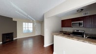 This single-floor space showcases an open-concept layout that seamlessly connects the kitchen and living area, creating a cohesive and functional environment. The rich, dark hardwood flooring flows throughout, providing a warm and inviting foundation that contrasts elegantly with the neutral, light-toned walls.

The kitchen is positioned along one side of the space and features dark mahogany-toned cabinetry that provides ample storage. The countertops are finished in granite with natural variation in tone, adding texture and visual interest. A peninsula-style breakfast bar extends outward, offering a casual dining or workspace option while maintaining visual openness. Stainless steel appliances, including a microwave and electric range, are integrated into the kitchen design.

The adjoining living area benefits from natural light streaming in through a large window, brightening the space and offering views of the surrounding greenery. A textured ceiling adds subtle dimension overhead. The front entry door is positioned near the living area, making for a practical and straightforward layout. Overall, the space presents a clean, well-maintained interior with a balanced combination of functional design and comfortable finishes.
