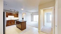 This single-floor residence offers a thoughtfully designed open-concept layout that seamlessly connects the kitchen, dining, and living areas. The kitchen is outfitted with rich dark wood cabinetry, a central island with a dark countertop providing additional prep and storage space, tile flooring, and stainless steel appliances. Track lighting overhead adds both function and a modern touch to the culinary space.

The living area transitions smoothly from tile to plush carpeting, creating a warm and inviting atmosphere. A tray ceiling adds architectural interest and a sense of elevated space to the main living zone. Large sliding glass doors flood the room with natural light and open to a private balcony, offering a connection to the outdoors.

A hallway extends from the main living area, leading to additional rooms and providing a clear and logical flow throughout the residence. Neutral tones in warm beige and soft gray are carried throughout, contributing to a clean, cohesive aesthetic. Recessed lighting complements the natural light and ensures the space feels bright and welcoming at all hours. Overall, the layout balances open communal living with defined private spaces, making it both practical and comfortable.