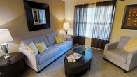 This single-floor space centers around a well-appointed living room that strikes a balance between comfort and style. The room is anchored by a light gray sofa accented with yellow and white patterned throw pillows, creating a warm, inviting atmosphere. A dark oval coffee table serves as the focal point of the seating area, complementing the rich, dark-framed wall mirror that adds depth to the neutral-toned walls.

Natural light filters into the space through a large window dressed with dark brown sheer curtains over white horizontal blinds, offering both ambiance and privacy control. A classic table lamp on a side console provides warm supplemental lighting, enhancing the cozy feel of the room.

The carpeted flooring contributes to the space's comfortable and relaxed character, while the neutral wall color provides a versatile backdrop for the furnishings. The overall design palette — combining warm taupes, deep browns, and accents of yellow — creates a cohesive and welcoming environment. The layout is practical and uncluttered, making efficient use of the available space while maintaining an open, livable feel.