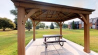 This outdoor pavilion is a well-constructed, single-level open-air structure built with sturdy timber framing. The gabled roof features exposed wooden beams and rafters arranged in a classic ridge pattern, topped with a corrugated metal covering that provides reliable shelter from the elements. Substantial square wooden posts support the structure at each corner, lending it a solid, durable character while maintaining an open, airy feel on all sides.

The pavilion's concrete floor provides a clean, level surface underfoot, and a metal picnic table with bench seating is positioned centrally beneath the roof, offering a functional gathering space for outdoor dining or relaxation. The open design ensures unobstructed sightlines and natural airflow throughout.

Surrounding the pavilion is a well-maintained grassy landscape with mature trees, including evergreens and broadleaf varieties, that frame the views from beneath the structure. A nearby building with balconies is visible in the background, suggesting the pavilion is part of a larger community or residential property. The overall setting is peaceful and park-like, making this pavilion a practical and attractive amenity for outdoor gatherings in a natural environment.