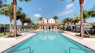 This outdoor amenity space showcases a striking resort-style pool environment designed with a Mediterranean architectural influence. The centerpiece is a generously sized, crystal-clear swimming pool with a rectangular layout, flanked on both sides by wide, light-colored concrete decking that provides ample space for lounging. Rows of chaise lounge chairs line each side of the pool, offering residents or guests comfortable spots to relax under the Florida sun.

At the far end of the pool, a covered pavilion with a terracotta-tiled roof serves as a shaded gathering area, seamlessly connecting the aquatic space to the surrounding buildings. The backdrop features a multi-story white stucco building with large windows and balconies, consistent with a Mediterranean or Spanish Colonial design aesthetic. Tall, mature palm trees are strategically placed throughout the space, providing natural shade and enhancing the tropical ambiance.

On either side of the pool area, elegant private cabanas with neutral-toned canopies offer more secluded relaxation options. The overall atmosphere is polished and resort-like, combining lush tropical landscaping with clean architectural lines to create an inviting and upscale outdoor retreat.