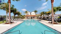 This outdoor amenity space showcases a striking resort-style pool environment designed with a Mediterranean architectural influence. The centerpiece is a generously sized, crystal-clear swimming pool with a rectangular layout, flanked on both sides by wide, light-colored concrete decking that provides ample space for lounging. Rows of chaise lounge chairs line each side of the pool, offering residents or guests comfortable spots to relax under the Florida sun.

At the far end of the pool, a covered pavilion with a terracotta-tiled roof serves as a shaded gathering area, seamlessly connecting the aquatic space to the surrounding buildings. The backdrop features a multi-story white stucco building with large windows and balconies, consistent with a Mediterranean or Spanish Colonial design aesthetic. Tall, mature palm trees are strategically placed throughout the space, providing natural shade and enhancing the tropical ambiance.

On either side of the pool area, elegant private cabanas with neutral-toned canopies offer more secluded relaxation options. The overall atmosphere is polished and resort-like, combining lush tropical landscaping with clean architectural lines to create an inviting and upscale outdoor retreat.