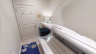 This compact, single-purpose tanning room is designed with functionality and comfort in mind. At its center sits a Sun Dash tanning bed, a full-length unit that serves as the focal point of the space. The room is finished with neutral, light-toned walls that create a clean and calming environment, complemented by warm ambient lighting provided by a small table lamp resting on a teal accent side table. The side table also offers a convenient surface for personal items or tanning accessories. A decorative blue rug with a tropical palm tree motif adds a touch of personality to the tiled floor, reinforcing a relaxed, resort-like atmosphere. A private entry door with a lock ensures privacy during use, and small hooks mounted near the door provide a practical spot for hanging clothing or accessories. A framed notice is displayed on the wall near the door, likely containing usage instructions or guidelines for the room. The space is efficiently laid out, ensuring ease of access to the tanning bed while maintaining a tidy, uncluttered feel. Overall, this tanning room delivers a private, well-appointed environment for indoor tanning.