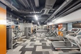Located at 4849 Connecticut Ave NW in Washington, DC, this well-appointed fitness center offers a comprehensive workout environment across a single open floor. The gym's industrial-inspired design is immediately evident through its exposed ceiling, where dark-painted ductwork and overhead piping lend a contemporary, urban aesthetic. Bright linear lighting runs the length of the space, ensuring consistent illumination throughout. The flooring features a bold geometric patterned carpet in varying shades of grey, adding visual interest while defining different workout zones. Along one side, a row of Star Trac treadmills and elliptical machines provides ample cardio options, while a dedicated section of strength and resistance equipment occupies the opposing area. Large wall mirrors amplify the sense of space and allow members to monitor their form during exercise. A designated stretching and floor exercise zone is equipped with exercise mats and free weights, offering a comfortable area for warm-ups and cool-downs. Orange accent walls punctuate the grey-toned interior, injecting energy and vibrancy into the space. A water station and waste disposal units are conveniently positioned for member use. The layout is thoughtfully organized to allow smooth circulation between workout areas, making this fitness center a functional and motivating environment for residents and users alike.