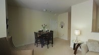 This single-floor space presents a well-arranged dining area as its focal point, featuring a dark wood high-top table paired with four matching bar-height chairs. The table is accented with a floral centerpiece and elegant candlestick holders, adding a decorative touch to the space. The dining area is set atop neutral-toned carpeting that extends throughout, contributing to a warm and inviting atmosphere.

The walls are finished in a soft, light yellow-green hue, lending a calm and understated character to the interior. A decorative wall art piece depicting a stylized tree adds a modest artistic element to the otherwise minimal décor. A standard white paneled door is positioned along one wall, providing access to an adjoining area.

Overhead, a flush-mounted ceiling light provides ambient illumination, while a table lamp on a glass-topped side table near the edge of the space offers additional warm lighting. The open layout allows for easy transition between the dining area and the adjacent living space, where the corner of a chair is visible, suggesting a comfortable and functional floor plan throughout.