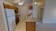 This single-floor space centers around a practical, well-equipped kitchen that serves as the heart of the layout. The kitchen features warm honey-toned wood cabinetry providing ample upper and lower storage, complemented by neutral laminate countertops. White appliances include a coil-top electric range, dishwasher, refrigerator, and an over-the-counter microwave, offering a complete and functional cooking setup. A peninsula breakfast bar extends from the main counter, creating a natural division between the kitchen and the adjoining living area while providing additional prep or casual dining space. Light ceramic tile flooring covers the kitchen zone, offering durability and ease of maintenance, while plush carpet transitions into the surrounding living spaces. The vaulted ceiling adds a sense of openness and volume to the space, enhanced by a ceiling fan with integrated lighting that ensures both comfort and illumination. A hallway visible beyond the kitchen leads to additional rooms, suggesting a practical and connected floor plan throughout the unit.