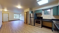 This single-floor unit offers a functional and well-appointed layout with clearly defined living, kitchen, sleeping, and bathroom areas. The open living space is finished with light wood laminate flooring and neutral walls, creating a bright and clean atmosphere. Corner windows allow natural light to fill the room, providing views of the surrounding greenery.

The kitchen is a standout feature, equipped with teal-toned cabinetry that extends to the ceiling, dark granite-style countertops, a stainless steel refrigerator, electric range, and dishwasher. A skylight overhead adds additional natural illumination to the cooking space, while a side window further enhances ventilation and light. Ample upper and lower cabinetry provides generous storage throughout.

Adjacent to the kitchen, a built-in closet with wood-panel sliding doors offers convenient storage. The bedroom is carpeted in a neutral blue-grey tone and features a window with an outdoor view, along with baseboard heating. The bathroom includes a full soaking tub with grab bars for accessibility and is positioned alongside a stacked in-unit washer and dryer. The overall design is practical and accessible, with consistent finishes throughout.