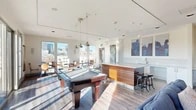 Located at 1756 N Clarkson St in Denver's Capitol Hill neighborhood, this contemporary amenity lounge is a well-appointed communal space designed for both recreation and relaxation. The open-plan layout is bathed in natural light, courtesy of expansive floor-to-ceiling windows and a sliding glass door that opens to an outdoor terrace, framing impressive views of the Denver skyline and surrounding cityscape.

The space is anchored by a full-size pool table positioned beneath a trio of industrial-style pendant lights, lending a stylish focal point to the room. Adjacent to it, a substantial wood-paneled wet bar with bar stool seating and a sink provides a functional entertaining hub, complemented by bold urban artwork displayed on the crisp white walls above.

A dedicated lounge area features comfortable blue upholstered seating arranged around a coffee table, while a separate sitting zone showcases a vibrant terracotta accent wall with a wall-mounted television and eclectic furnishings. Near the panoramic windows, bistro-style tables and chairs offer a bright spot for casual seating with sweeping city views. Light wood-tone flooring runs throughout, tying the space together with a clean, modern aesthetic. The terrace access further extends the usable space outdoors, making this lounge a versatile and inviting retreat.