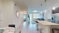 This single-floor residence at 2101 S Haverhill Rd in West Palm Beach showcases a contemporary design defined by clean lines, light-toned luxury vinyl flooring, and a bright, neutral color palette throughout. The open-concept layout connects the kitchen, dining, and living areas seamlessly, creating an airy and cohesive flow.

The kitchen is a standout feature, equipped with stainless steel appliances, dark espresso flat-panel cabinetry extending to ceiling height, white quartz countertops, a subway tile backsplash, and a central island with bar seating and pendant lighting overhead. The space is both functional and visually striking.

The living room receives generous natural light through multiple windows fitted with roller shades, and a sliding glass door provides direct access to an outdoor area with views of the surrounding landscape. The bedroom features a closet and direct access to a well-finished bathroom, which includes a single undermount sink with quartz countertops and a glass-enclosed shower.

Throughout the unit, recessed lighting and pendant fixtures complement the natural light, reinforcing the modern, polished ambiance. The consistent use of light flooring, white walls, and contemporary fixtures gives the entire space a cohesive, move-in-ready aesthetic.