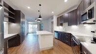 This contemporary single-floor residence offers a thoughtfully designed open-concept layout that seamlessly connects the kitchen and living areas. The kitchen is a standout feature, equipped with dark espresso cabinetry accented by sleek brushed nickel hardware, a crisp white quartz countertop, and a large center island ideal for dining or meal prep. Stainless steel appliances, including a dishwasher, range, and microwave, complement the space, while a white subway tile backsplash and under-cabinet lighting add a refined touch. Stylish industrial-style pendant lights hang above the island, enhancing the modern aesthetic. Warm hardwood-style flooring flows throughout the main living areas, while a ceiling fan provides comfort. Large windows and a glass-door balcony access flood the living space with natural light and provide an outdoor retreat. The bedroom features plush carpeting, a ceiling fan, and generous wall space. The bathroom is appointed with a white quartz vanity, dark cabinetry, and open built-in shelving, offering both style and practical storage. Additional built-in shelving and tall pantry-style cabinetry in the kitchen area provide ample storage throughout this well-appointed residence.