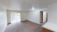 This single-floor unit offers two bedrooms and one bathroom within a well-proportioned floor plan. The living area serves as the central gathering space, featuring wall-to-wall carpeting in a neutral tone that adds warmth underfoot. A large sliding glass door fitted with vertical blinds allows natural light to fill the room while providing direct access to an outdoor area, creating a bright and open atmosphere.

The ceiling features a textured finish, and flush-mount lighting provides consistent illumination throughout. Neutral white walls throughout the living space offer a clean, versatile backdrop. A floor-level vent indicates a forced-air heating and cooling system for year-round comfort. Wood-toned baseboards frame the carpeted floor, adding a subtle contrast to the light walls.

A partial view of cabinetry in the foreground suggests the kitchen is positioned adjacent to the living area, supporting an open and connected layout. A hallway visible at the rear of the living space leads to the bedrooms and bathroom, providing a clear and functional traffic flow. The overall design emphasizes simplicity, practicality, and comfortable everyday living.