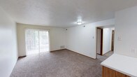 This single-floor unit offers two bedrooms and one bathroom within a well-proportioned floor plan. The living area serves as the central gathering space, featuring wall-to-wall carpeting in a neutral tone that adds warmth underfoot. A large sliding glass door fitted with vertical blinds allows natural light to fill the room while providing direct access to an outdoor area, creating a bright and open atmosphere.

The ceiling features a textured finish, and flush-mount lighting provides consistent illumination throughout. Neutral white walls throughout the living space offer a clean, versatile backdrop. A floor-level vent indicates a forced-air heating and cooling system for year-round comfort. Wood-toned baseboards frame the carpeted floor, adding a subtle contrast to the light walls.

A partial view of cabinetry in the foreground suggests the kitchen is positioned adjacent to the living area, supporting an open and connected layout. A hallway visible at the rear of the living space leads to the bedrooms and bathroom, providing a clear and functional traffic flow. The overall design emphasizes simplicity, practicality, and comfortable everyday living.