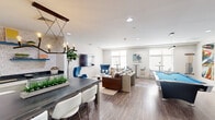 Located at 240 Sidney St in Cambridge, MA, this contemporary community room offers a well-designed, multi-functional gathering space spread across a single floor. The room is finished with warm wood-look flooring, recessed lighting, and a bright, neutral color palette that lends a clean, modern aesthetic throughout.

The space is organized into several distinct activity zones. A central pool table anchors the recreational area, accompanied by a bold, large-scale abstract artwork that adds a vibrant focal point. Adjacent to it, a comfortable lounge area features a leather sofa, distinctive blue egg-style chairs, and a wall-mounted flat-screen TV set against subtly patterned wallpaper. A kitchenette with dark countertops and open shelving supports the dining area, which includes a long communal table with mixed seating.

High-top bar tables and bar-stool seating line the window-facing perimeter, allowing for ample natural light. A secondary lounge nook near the entrance features plush yellow accent chairs, a marble-top side table, and a decorative geometric wall panel alongside glass entry doors. A compact workspace with a wall-mounted TV and a built-in desk rounds out the room's offerings. Together, these zones create a versatile, inviting environment well-suited for socializing, dining, and recreation.