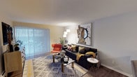 This single-floor space offers a thoughtfully designed living area that balances comfort and contemporary style. The living room serves as the focal point of the space, anchored by a dark sectional sofa accented with an array of decorative throw pillows in gold, charcoal, and patterned tones, complemented by a cozy mustard-yellow throw blanket. A bold, oversized abstract artwork commands attention on the main wall, adding a dramatic visual element to the room. An accent chair in a vibrant orange-red hue introduces a pop of color, while a multi-tiered floor lamp provides warm ambient lighting. The flooring features a geometric-patterned area rug in neutral tones with yellow accents, layered over what appears to be carpeted flooring. A glass-top coffee table and a sleek side table round out the seating arrangement. Large windows fitted with vertical blinds allow abundant natural light to filter into the space, creating a bright and airy atmosphere. The neutral wall palette in soft whites and warm beige tones provides a versatile backdrop that enhances the room's modern aesthetic. Overall, the space presents a well-curated, inviting environment suitable for both relaxing and entertaining.