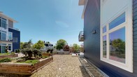 Located at 30 Haven St in Reading, Massachusetts, this single-floor exterior space offers a well-appointed outdoor environment that combines functional design with thoughtful landscaping. The building itself features a striking dark navy horizontal lap siding exterior, lending a modern and contemporary aesthetic to the property.

At ground level, a light-toned composite deck runs along the building's facade, providing a clean and low-maintenance outdoor surface. Large sliding glass doors open directly from the deck to the interior, allowing for seamless indoor-outdoor connectivity and abundant natural light to flood the adjacent interior spaces. Windows along the exterior reveal a fitness center inside, equipped with cardio machines, reflecting the building's commitment to resident amenities.

The surrounding yard features a gravel ground cover that creates a tidy, permeable landscape, bordered by rustic wooden raised garden beds that add a touch of greenery and a practical gardening element to the outdoor area. A patio area with fencing is visible further in the yard, suggesting a designated gathering or relaxation space. Mature trees from the neighboring properties contribute to a pleasant, leafy backdrop.

The space enjoys excellent natural light under open skies, situated within a well-established residential neighborhood. Overall, the exterior presents a cohesive blend of modern architectural details and practical outdoor living features.