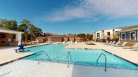 Located at 3761 Raleigh Road Parkway in Wilson, North Carolina, this single-level outdoor amenity space is designed to deliver a resort-style experience for its community. At the heart of the space is a generously sized swimming pool with crystal-clear water, flanked by a wide concrete pool deck lined with lounge chairs and shaded cabanas, ideal for relaxing in the North Carolina sunshine.

A standout feature is the covered pavilion, which is anchored by striking stacked-stone columns and finished with a warm wood-paneled ceiling. The pavilion serves as a versatile outdoor living area, equipped with a wall-mounted flat-screen television and a sleek linear fireplace set into a classic brick surround — perfect for year-round enjoyment. Multiple seating arrangements, including round dining tables and chairs, provide comfortable spaces for gathering and entertaining.

The pavilion opens directly toward the pool deck, creating a seamless indoor-outdoor flow. Views from the covered area extend across well-maintained green lawns and lush mature trees, framed by decorative iron fencing. The thoughtful combination of natural materials — stone, wood, and brick — alongside modern amenities gives this outdoor space a polished yet welcoming ambiance suited for both relaxation and social gatherings.