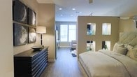 This single-floor studio is designed with a smart, open layout that maximizes both functionality and style. A defining architectural feature is the partial room divider wall, which cleverly separates the sleeping area from the living space without fully enclosing either zone. The divider is enhanced with open cutout niches, providing decorative display space for decorative accents such as vases and glassware, adding a touch of personality and design cohesion throughout.

The sleeping area is anchored by a platform-style bed dressed in crisp white linens, positioned alongside a bold dark-stained dresser topped with a classic table lamp and decorative bowl. Warm taupe walls and rich hardwood-style flooring run continuously throughout, lending a cohesive and grounded aesthetic to the entire space.

The living area beyond the divider benefits from generous natural light streaming through large windows with horizontal blinds, illuminating a neutral-toned seating arrangement and a cowhide-style area rug. Recessed lighting in the white ceiling complements the natural light and ensures the space remains well-lit at all times. The overall ambiance is modern and polished, with a warm, neutral palette that creates a welcoming and refined atmosphere throughout.