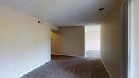 This single-floor space presents a straightforward, functional layout with an open living area that connects to an adjoining room, offering a sense of continuity throughout. The flooring throughout is covered in dark wall-to-wall carpeting, providing a soft, uniform foundation across the space. Walls are finished in a neutral beige tone, creating a clean and understated backdrop well-suited for a variety of interior styles.

The ceiling features a textured popcorn finish and includes multiple HVAC vents, ensuring climate control across the space. White baseboards run along the perimeter of the walls, offering a subtle contrast to the beige tones. A partial wall divider separates the main living area from the adjoining room, defining distinct zones while maintaining an open feel.

The adjoining room, visible through the open passage, mirrors the same carpeting and wall tones, reinforcing the cohesive aesthetic of the space. The layout appears efficient, with clear sightlines between areas. Overall, the space offers a neutral, move-in-ready environment with a practical single-level design.