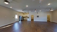 This single-story space offers an open-concept layout that seamlessly integrates the living area and kitchen into one expansive, unified environment. The rich, dark hardwood-style flooring runs throughout, providing a warm and grounded aesthetic that contrasts cleanly with the bright white walls and neutral-toned ceilings.

The kitchen is positioned along the back wall and features white cabinetry, stone or granite-look countertops, stainless steel appliances including a full-size refrigerator, and a practical layout with ample workspace. Recessed lighting above the kitchen ensures the area is well-lit for functional use.

The open living area is notably spacious, offering flexibility for various furniture arrangements. Two ceiling fans with integrated light fixtures are evenly distributed across the ceiling, contributing both to air circulation and ambient lighting. Recessed lighting is also installed throughout, supplementing natural light that enters through a side door with a window panel.

Crown molding details along the upper walls add a subtle architectural refinement to the space. A secondary interior door is visible toward the rear, suggesting additional rooms or utility areas beyond the main open space.