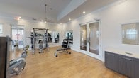 Located at 1500 Water Lily Dr in Simpsonville, SC, this single-floor fitness center is thoughtfully designed to support a comprehensive workout experience. The space features warm hardwood flooring throughout, crisp white walls, and recessed lighting complemented by ceiling fans that maintain airflow and comfort during workouts.

The facility is divided into distinct functional zones. The cardio area is outfitted with multiple True-brand treadmills, an elliptical trainer, a recumbent bike, a spin bike, and a rowing machine — all positioned near large windows fitted with white blinds that allow ample natural light to fill the room. Wall-mounted flat-screen televisions are positioned throughout to enhance the workout experience.

The strength training zone features a multi-functional cable machine, a Smith machine, a dedicated dumbbell rack with a full selection of free weights, and an adjustable workout bench. Full-length mirrors line the walls in the strength area, creating a sense of expanded space while allowing proper form checks during lifts.

The space is accessed through elegant glass-paned French doors, adding a refined architectural touch to the otherwise functional environment. The open floor plan provides generous room to move freely between equipment, making this fitness center both practical and inviting for daily use.