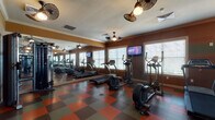 This fitness center is a thoughtfully designed, single-level exercise space offering a comprehensive range of equipment for both cardio and strength training. The room is finished with warm, earthy-toned walls accented by crown molding, and a textured ceiling equipped with multiple decorative ceiling fans that provide consistent air circulation throughout. The distinctive red and olive checkered rubber flooring adds a bold visual element while offering durable, cushioned support underfoot.

The cardio zone features multiple treadmills and elliptical trainers arranged along the windowed wall, where large windows fitted with horizontal blinds allow ample natural light to fill the space. Wall-mounted flat-screen televisions are positioned at eye level, providing entertainment during workouts. A full-length mirror along one wall visually expands the space and allows users to monitor their form.

On the opposite side of the room, a multi-station cable weight machine offers a variety of strength training options, complemented by additional cardio equipment including a stationary bike. A small utility shelf near the entryway provides convenient storage for fitness accessories. The space is accessed through a glass-panel door and includes clearly marked emergency exits, ensuring both functionality and safety.