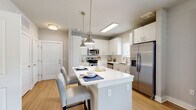 Located in St. Augustine, FL, this well-appointed single-floor residence offers a clean, modern aesthetic with thoughtful finishes throughout. The open-concept kitchen is a standout feature, showcasing white shaker cabinetry, a classic subway tile backsplash, stainless steel appliances, and a generous island with bar seating illuminated by polished pendant lights. Light wood-look flooring flows through the main living areas, adding warmth and continuity to the space. The living room is bright and inviting, with multiple large windows that frame outdoor greenery, allowing abundant natural light to fill the room. French doors provide additional access and further enhance the airy feel of the space. The bedrooms are carpeted for comfort and feature soft, neutral tones, with windows dressed in layered curtains that allow for both natural light and privacy. The bathroom is finished with white subway tile surrounding a full tub-and-shower combination, paired with a white vanity featuring modern brushed gold hardware and a clean countertop. A dedicated hallway area with light wood cabinetry and multiple interior doors provides practical storage and room separation. The overall ambiance of the residence is calm, contemporary, and well-lit, making it a comfortable and functional home.