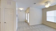 This single-floor space presents a clean, understated interior defined by neutral cream-toned walls and soft, light grey carpeting throughout. The open layout creates a natural flow between living areas, with a partially vaulted ceiling adding a sense of vertical spaciousness to the main room. A window allows natural light to filter in, brightening the interior and enhancing the airy feel of the space. HVAC vents are integrated into both the walls and ceilings, ensuring efficient climate control throughout. The layout includes a connecting hallway that leads to additional areas, with a ceiling-mounted light fixture providing ambient illumination. White trim and baseboards offer clean contrast against the warm neutral walls, contributing to a simple, cohesive aesthetic. The space is move-in ready with a blank-canvas quality, allowing for flexible furnishing and personalization. Overall, the interior maintains a modest, functional character well-suited for comfortable everyday living.