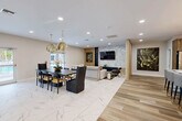 Located at 140 SW 91st Ave in Plantation, FL, this single-floor space spans a generously sized footprint and showcases a cohesive contemporary design throughout. The interior is unified by large-format marble-look porcelain tile and warm wood-grain tile flooring, creating a refined yet inviting atmosphere.

The grand reception area features soaring ceilings with a coffered tray detail, dramatic sputnik chandeliers, floor-to-ceiling sheer drapery, and expansive windows that flood the space with natural light. White marble-look flooring anchors this airy, open area, which flows seamlessly into the rest of the layout.

The kitchen is appointed with wood-grain cabinetry, gold hardware, a white quartz island with decorative tile detailing, bar seating, subway tile backsplash, and tiered gold pendant lighting. Stainless steel appliances and a wine cooler complete the functional design.

The open-plan living and dining area features a striking marble-clad accent wall with a mounted television, flanked by vertical wood slat panels and built-in shelving. A hallway with plantation shuttered storage connects the zones efficiently. A separate dining lounge offers curved booth seating in sage green with a tropical botanical mural, gold-accented pendant lighting, and a warm wood divider—an intimate setting within the broader space.