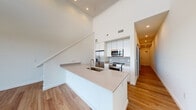 Located at 101 Manchester Rd in Richmond, Virginia, this contemporary two-floor residence offers a thoughtfully designed layout with a clean, modern aesthetic throughout. The main level centers around an open-concept living and kitchen area, where soaring double-height ceilings create a dramatic sense of volume and airiness. The sleek kitchen features white cabinetry extending to the ceiling, stainless steel appliances including a refrigerator, range, and microwave, and a large peninsula with a quartz countertop and integrated sink — ideal for both cooking and casual dining. Warm hardwood flooring flows seamlessly throughout the space, complementing the bright white walls and recessed lighting. A staircase connects the two levels, reinforcing the open, loft-like feel of the main living area. Sliding glass doors open to a private balcony, offering open views of the surrounding Richmond cityscape and filling the space with abundant natural light. The upper level provides well-proportioned rooms with continued hardwood flooring and a clean, minimalist finish. A hallway with louvered closet doors leads to a full bathroom, finished with light-toned tile flooring, a soaking tub, and a wood-accented vanity with a quartz countertop. The overall ambiance is bright, modern, and move-in ready.