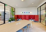 Located at 51 NW 28th St in Miami's creative Wynwood neighborhood, this thoughtfully designed workspace offers a refined yet vibrant environment ideal for professional collaboration and focused work. The single-floor layout is defined by sleek black-framed glass partition walls that divide the space into distinct functional zones while maintaining an open, airy feel throughout.

Light hardwood flooring runs continuously across the space, complementing the neutral white walls and crisp contemporary finishes. A large central work table with light wood surfaces anchors the main area, surrounded by modern stools and accent chairs that encourage both individual productivity and group interaction. Along one wall, a built-in low credenza with integrated cabinetry is topped by a floating display shelf styled with curated decorative objects, lending the space a polished, gallery-like quality.

A standout lounge zone features a row of bold red high-back booth seating with tufted upholstery, paired with colorful framed artwork above — adding a striking visual focal point. Lush tropical potted plants are positioned throughout, bringing organic warmth and vibrancy in keeping with Miami's character. Floor-to-ceiling windows along the exterior wall flood the space with natural light and offer expansive city views, creating a bright and energizing atmosphere throughout the day.