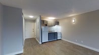 This single-floor unit offers a thoughtfully designed open-concept layout that efficiently combines living and kitchen areas. The kitchen is neatly integrated into the space, featuring dark navy lower cabinetry, gray upper cabinets, a white refrigerator, a compact range, and a single-basin sink — all arranged to maximize functionality within a cozy footprint. The kitchen's design creates a natural visual anchor within the open living area without feeling intrusive.

The flooring throughout is a warm, light-toned hardwood-style material that adds continuity and a sense of spaciousness to the interior. Neutral gray-toned walls complement the overall palette, creating a calm and modern ambiance. Flush-mounted ceiling lights provide consistent, even illumination, while a wall sconce near the kitchen adds a softer accent light.

A hallway leads from the main living area to the unit's entry door, which is fitted with a classic white panel design and standard hardware. The textured ceiling adds a subtle architectural detail while keeping the overall feel clean and understated. The space is well-maintained and move-in ready, offering a practical and comfortable living environment with a cohesive, contemporary design sensibility throughout.