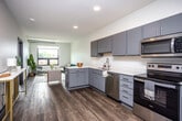 This single-floor unit offers a well-designed open-concept layout that seamlessly connects the kitchen and living areas, creating an efficient and inviting flow. The kitchen is outfitted with gray shaker-style cabinetry, white quartz countertops, and a full suite of stainless steel appliances, including a refrigerator, electric range, over-the-range microwave, and dishwasher. A peninsula with seating provides additional workspace and a casual dining option, while recessed lighting enhances the bright, clean aesthetic. Rich wood-style flooring runs throughout the entire unit, adding warmth and visual continuity.

The living area benefits from large windows that allow natural light to pour in and offer outdoor views of the surrounding neighborhood. Exposed ductwork along the ceiling adds an industrial design element to the space. Two bedrooms are included, each featuring the same wood-style flooring and large windows with exterior views, creating comfortable and naturally lit retreats.

The bathroom is finished with a gray vanity, white quartz countertop, an integrated sink, a large wall-mounted mirror, and a shower-tub combination. The overall design palette of gray, white, and warm wood tones gives the unit a modern and cohesive aesthetic throughout.