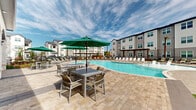 Located at 12202 Huffmeister Road in Cypress, Texas, this outdoor amenity space is designed to deliver a resort-style experience within a modern residential community. The centerpiece is a large, freeform swimming pool with crystal-clear blue water, surrounded by an expansive stone paver deck that provides ample room for lounging and socializing. Rows of poolside chaise lounges offer plenty of space for sunbathing, while a pergola structure at the far end of the pool adds a shaded retreat for relaxed gatherings.

Adjacent to the pool, a covered colonnade serves as an elegant transition between interior and exterior spaces. Defined by substantial cut-stone columns and arched architectural detailing, this covered area features large floor-to-ceiling glass windows that frame views of the pool deck, filling the interior lounge with natural light. Wicker seating with soft cushions provides a comfortable setting to take in the outdoor scenery.

The pool deck is outfitted with multiple dining tables paired with green market umbrellas, creating inviting spots for outdoor meals or casual conversation. Lush manicured turf borders portions of the deck, adding a touch of greenery to the hardscape. The surrounding multi-story residential buildings frame the space, reinforcing the community's cohesive and well-appointed architectural character. Overall, the space exudes a bright, open, and welcoming ambiance suited for year-round outdoor enjoyment.