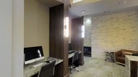 This single-floor space is a thoughtfully designed business center that blends modern aesthetics with functional workspaces. The interior is defined by a sophisticated palette of warm wood paneling, stone-textured accent walls, and neutral-toned carpeting, creating a professional yet inviting atmosphere.

The space is organized around individual workstations, each equipped with desktop computers and generous desk surfaces finished in a dark stone or granite-style material. Structural columns clad in rich dark wood paneling divide the area into semi-private zones, offering a degree of separation between workstations while maintaining an open, connected feel.

Lighting plays a key role in the ambiance, with sleek vertical wall sconces mounted on the wood columns providing warm, focused illumination. These fixtures complement the recessed ceiling lighting, ensuring the entire space is well-lit for detailed work.

The accent wall, finished in a stacked natural stone veneer, adds a striking textural contrast to the smooth wood and neutral surfaces throughout. Ergonomic adjustable chairs accompany each workstation, reflecting the space's commitment to comfort and practicality.

Overall, this business center communicates a clean, upscale design sensibility, balancing purposeful functionality with refined architectural details to support a focused and professional environment.