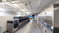 This single-floor shared laundry facility is designed to accommodate high-volume use while maintaining a clean, organized, and functional environment. The main laundry area is lined on both sides with rows of stainless steel commercial-grade front-loading washing machines and stacked dryers, all featuring modern digital controls. The polished concrete floors and white-painted ceiling with exposed ductwork and piping give the space an industrial yet tidy character, while overhead fluorescent lighting ensures the area is well-lit throughout.

A standout design feature is the reclaimed wood accent wall near the entrance area, which adds warmth and texture to an otherwise utilitarian setting. This transitions into a comfortable waiting lounge furnished with a sleek black sofa, mid-century modern chairs, and a wooden table set atop a bright yellow geometric-patterned rug. A large, vivid mural depicting a coastal sunset scene adorns one wall of the lounge, creating a vibrant focal point that brings color and energy to the space.

Small ground-level windows allow for a modest amount of natural light, complementing the artificial lighting throughout. A utility door provides access to a back service area, and informational postings are neatly displayed near the machines. Overall, this laundry facility combines practicality with thoughtful design touches that elevate the everyday laundry experience.