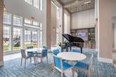 Located at 1700 CR-50 in Bay Shore, NY, this grand single-floor lobby and hallway space offers an impressive and welcoming environment defined by its soaring double-height ceilings and sweeping architectural scale. Large, floor-to-ceiling windows and transom glazing flood the interior with abundant natural light while providing views of the surrounding outdoor grounds. The expansive lobby features large-format white tile flooring that transitions into plush, patterned blue-toned carpeting in the seating and activity areas, adding warmth and visual interest throughout. A striking grand piano serves as a central focal point, positioned beneath the dramatic ceiling height and complemented by a built-in display niche. Circular dining tables paired with light wood and aqua upholstered chairs create inviting gathering spaces throughout the open floor plan. A stone-clad fireplace with a mounted television anchors one seating lounge, where bold navy sofas and accent chairs offer a comfortable retreat. A broad corridor lined with tall white columns extends from the main lobby, offering carpeted walkways illuminated by pendant lighting. Glass-paned exterior doors connect the interior to outdoor amenity spaces, reinforcing the bright, airy ambiance that defines this elegant and well-appointed communal facility.