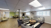 Located at 240 Village Crossing Ln in Winston-Salem, NC, this single-floor community space offers a thoughtfully designed, open layout that seamlessly connects multiple functional zones. The main lounge area is furnished with comfortable gray sofas and accent chairs arranged around a rustic wooden coffee table, set atop a bold black-and-white striped area rug. Natural light filters in through plantation-shuttered windows, complementing the warm wood-look flooring that runs throughout the space. Decorative artwork adorns the neutral-toned walls, contributing to an inviting and relaxed ambiance. Adjacent to the lounge, a dedicated dining area features a live-edge wooden table with ample seating, set against a vibrant green accent wall that adds a lively pop of color. A well-stocked bookcase and a cozy reading chair occupy a quiet corner, providing a more intimate retreat within the open floor plan. The fully equipped kitchen is outfitted with crisp white shaker cabinets, warm countertops, stainless steel appliances including a refrigerator, dishwasher, range, and built-in microwave, plus a double sink. A breakfast bar with bar seating provides an additional casual dining option. A drop ceiling with flush panel lighting illuminates the entire space evenly, creating a bright and welcoming environment throughout.