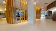 This single-floor space presents a notable contrast between two distinct areas. The lobby area is architecturally compelling, featuring a series of tall, sculpted golden wood slat installations that curve organically along the walls, creating a warm, textural focal point throughout the corridor. Large-format light-colored tile flooring extends across the space, reflecting the recessed overhead lighting and contributing to an open, polished ambiance. A glass-partitioned workspace is visible behind vertical wooden screens, housing display monitors and workstations. The reception area showcases a backlit, white marble-finish desk positioned beneath a bold, abstract sculptural wall piece in deep red tones. Floor-to-ceiling glass walls and bronze-framed entry doors allow natural light to pour in, connecting the interior to the outdoors. In contrast, the bathroom is modestly appointed with warm beige walls, an oak-cabinet vanity topped with a white basin and chrome fixtures, a framed mirror, and a standard toilet. White interior doors with brass hardware complete the functional, understated character of this secondary area.