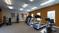 This single-floor fitness center is designed to provide a functional and comfortable environment for exercise. The space is finished with warm-toned accent walls in a rich amber hue, contrasting with crisp white ceilings and trim, creating an energizing atmosphere. Fluorescent lighting fixtures are evenly distributed across the ceiling, ensuring the room is well-lit throughout. Natural light also filters in through shuttered windows, adding to the bright and welcoming ambiance. The flooring is covered in durable, low-pile carpet suited for a fitness setting. The cardio area is well-appointed with multiple treadmills and an elliptical trainer, all positioned to take advantage of the natural light from the windows. A multi-function strength training machine is also present, offering users a variety of resistance exercises. A workout reference chart mounted on the wall provides additional guidance for users. An adjacent room is visible through an open doorway, suggesting additional support space such as a restroom or storage area. Overall, the fitness center is thoughtfully laid out to maximize usability, offering a range of equipment within a clean, organized, and inviting space.